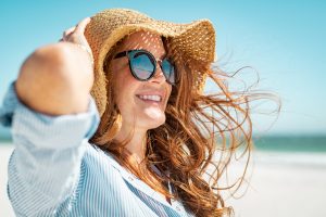 Side view of a women with long hair blowing in the wind wearing sunglasses and a straw hat at the beach. 