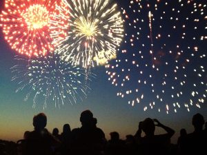 A Fourth of July fireworks display exploding in the sky over a group of people. The crowd, spectators in silhouette, celebrate the Independence Day national holiday together, looking up and watching bursts of color.