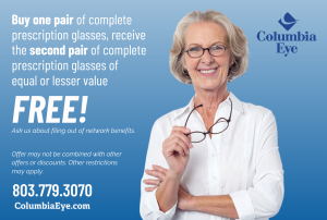 A smiling woman with short gray hair, wearing dark-framed glasses and a white blouse, holds a second pair of glasses in her hand. Buy one pair of complete prescription glasses, get a second pair of equal or lesser value free.